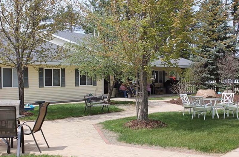 Outdoor garden area at Cedar Ridge Elder Services II with paved walkways, green grass, several trees, benches, and outdoor seating including tables and chairs. The building exterior is visible with light-colored siding and windows.