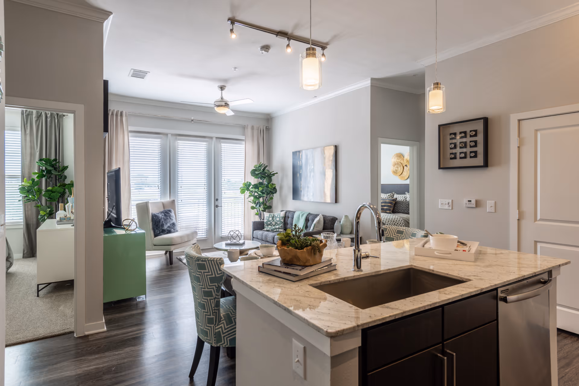 Modern open-concept living space with a kitchen island featuring a sink and countertop decor, a dining area with patterned chairs, a living room with a sofa, armchair, coffee table, and wall art, and a bedroom visible through an open doorway. Large windows with blinds and curtains allow natural light to fill the room.