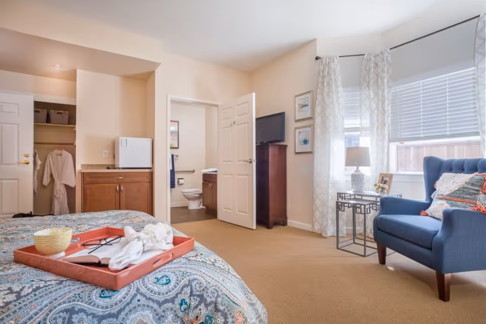 A cozy bedroom with a patterned bedspread and a red tray holding a bowl, an open book, and glasses. To the right, there is a blue armchair with decorative pillows next to a small side table with a lamp and a framed photo. The room has beige walls, a window with white blinds and patterned curtains, and an open door leading to a bathroom with a visible toilet and sink. There is also a closet with robes hanging inside and a small cabinet with a mini fridge on top.