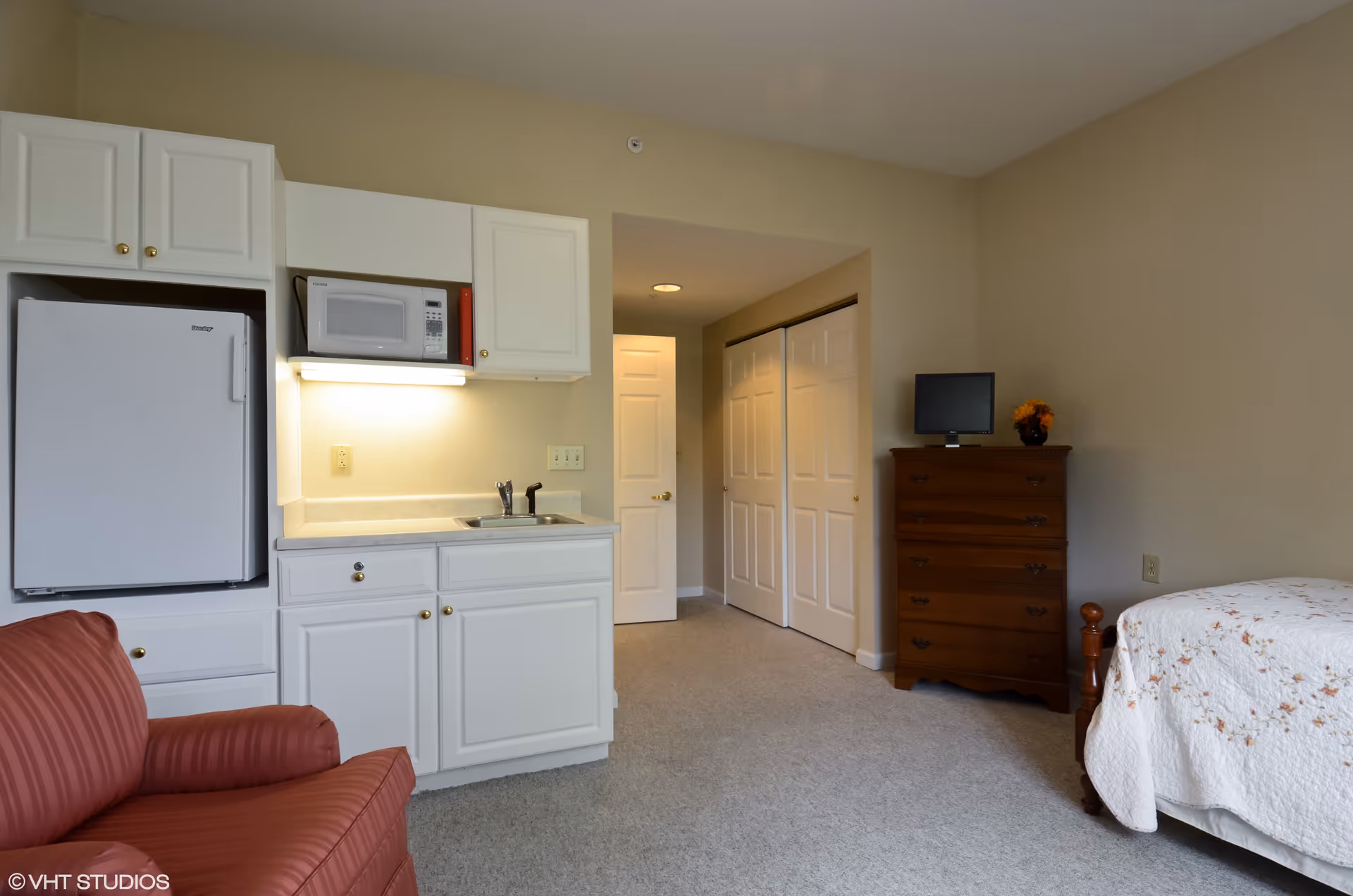 Interior view of a senior living facility room featuring a small kitchenette with white cabinets, a microwave, and a refrigerator. To the left is a red armchair, and to the right is a wooden dresser with a small TV and a flower vase on top. A bed with a white floral quilt is partially visible on the right side. The room has beige walls and carpeted flooring, with a hallway and closet doors in the background.