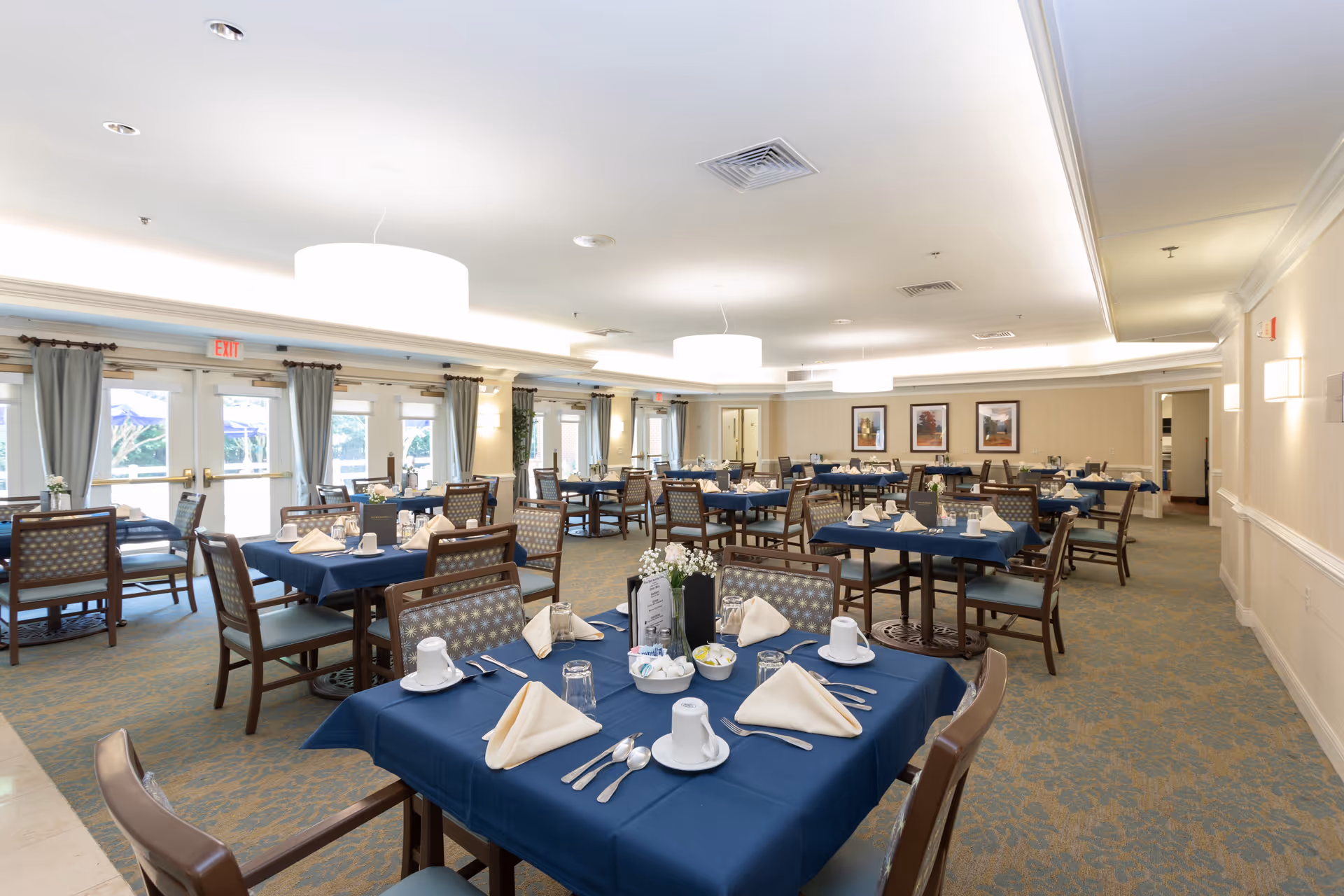 A spacious dining room with multiple tables covered in blue tablecloths, each set with white napkins, cups, glasses, and silverware. The room has large windows with curtains, soft lighting from ceiling fixtures, and framed artwork on the walls.