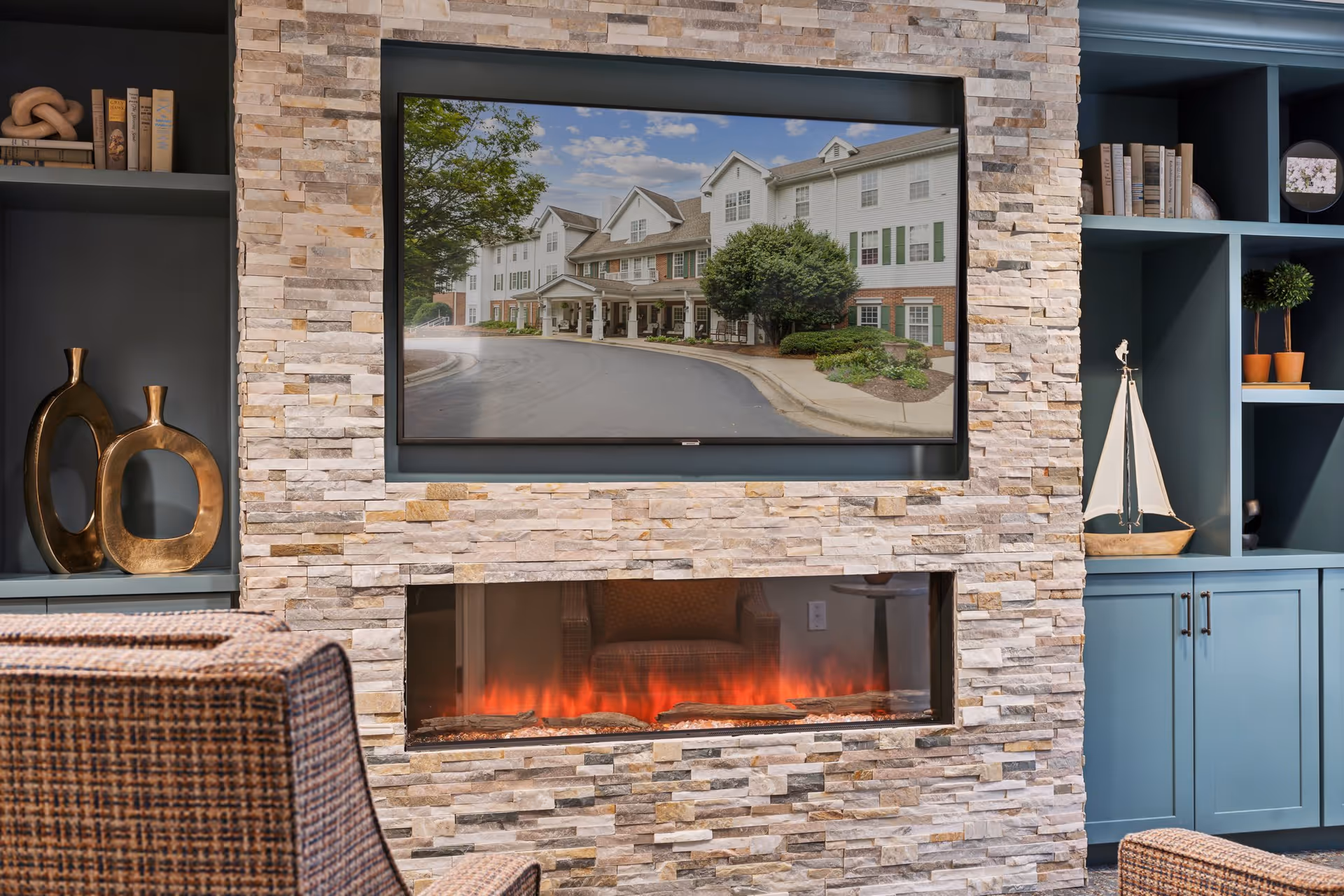 Cozy lounge with a stone-clad wall featuring a horizontal electric fireplace, a TV above it showing a building exterior, built-in blue shelving and seating.