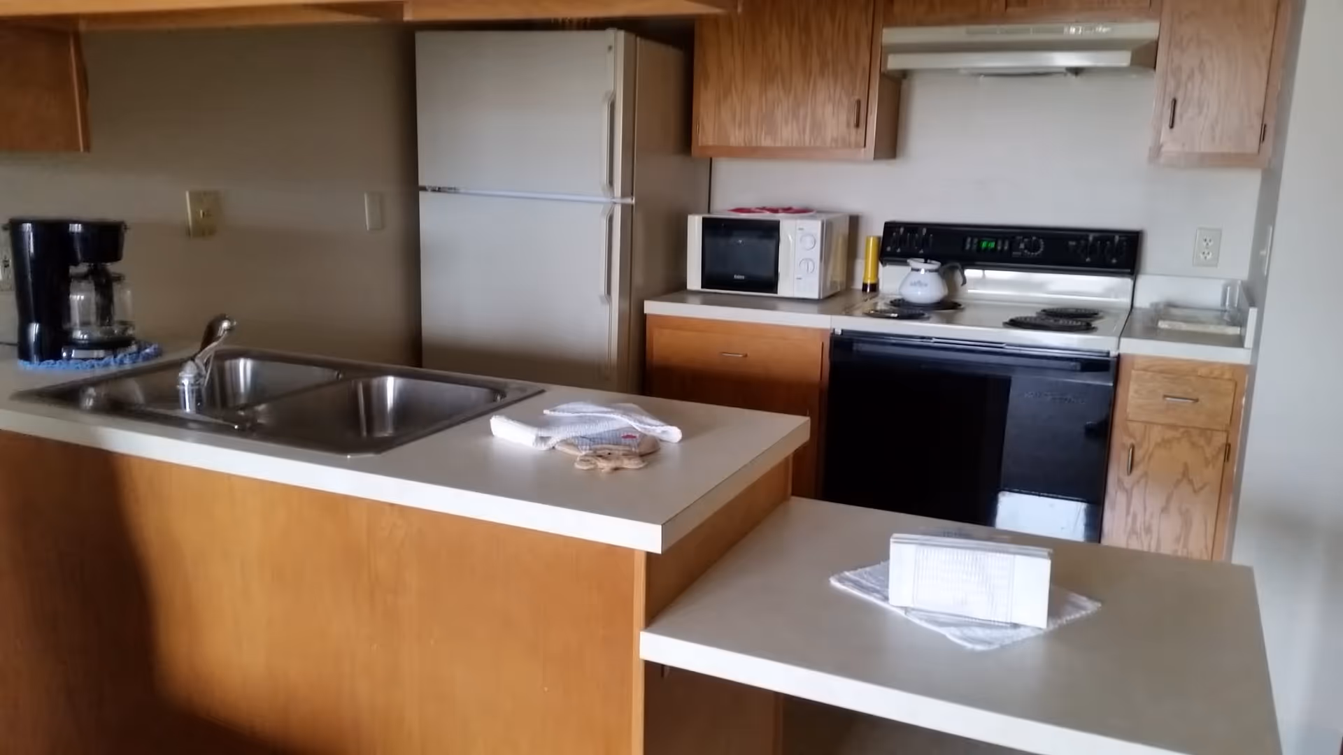 A kitchen area with wooden cabinets, a white refrigerator, a microwave, a black stove with a white kettle on it, a double sink, a coffee maker, and countertops with towels and a small item on them.