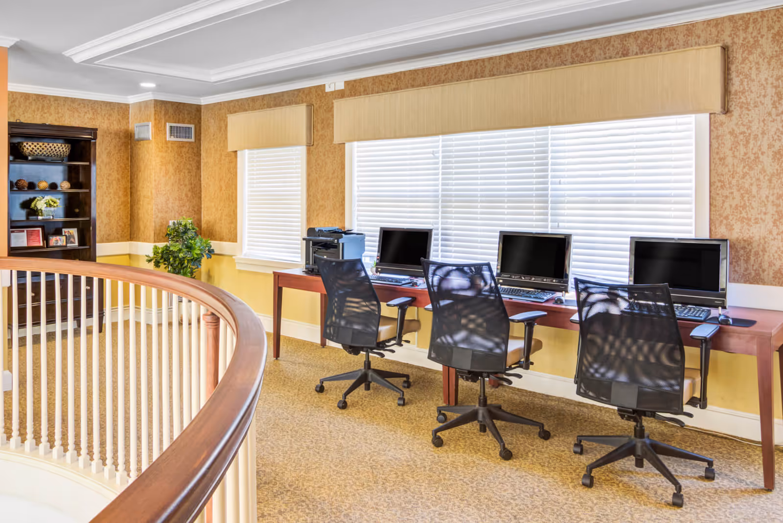 A bright communal computer workstation area with four desks and swivel chairs along windows and a curved wooden railing in the foreground.