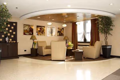 Cozy senior living lounge with beige chairs, side tables, lamps, artwork, and potted plants.