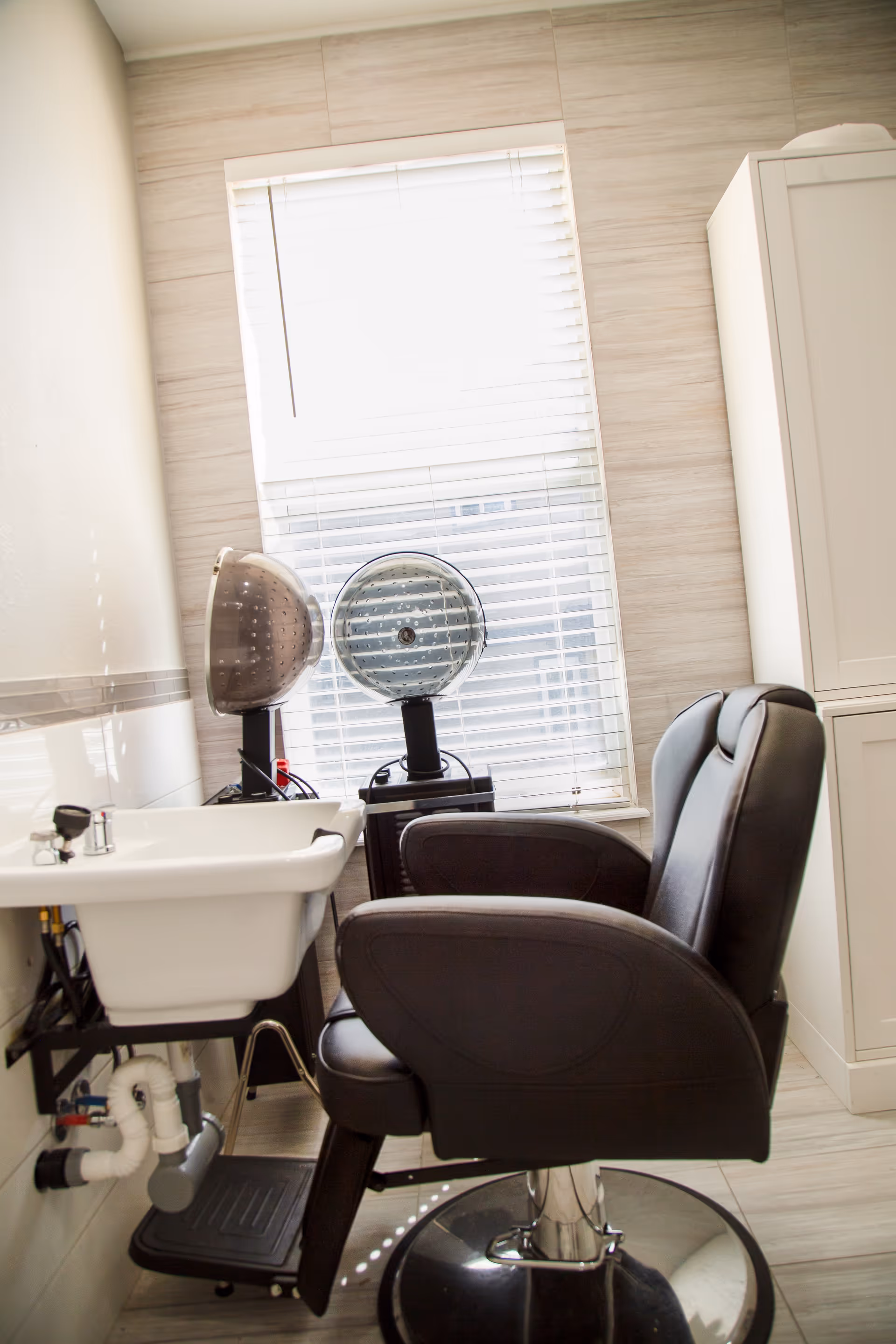 Interior view of a salon area in an assisted living facility featuring a black salon chair, a white sink with plumbing underneath, two hair dryer hoods, a window with white blinds, and a white cabinet.