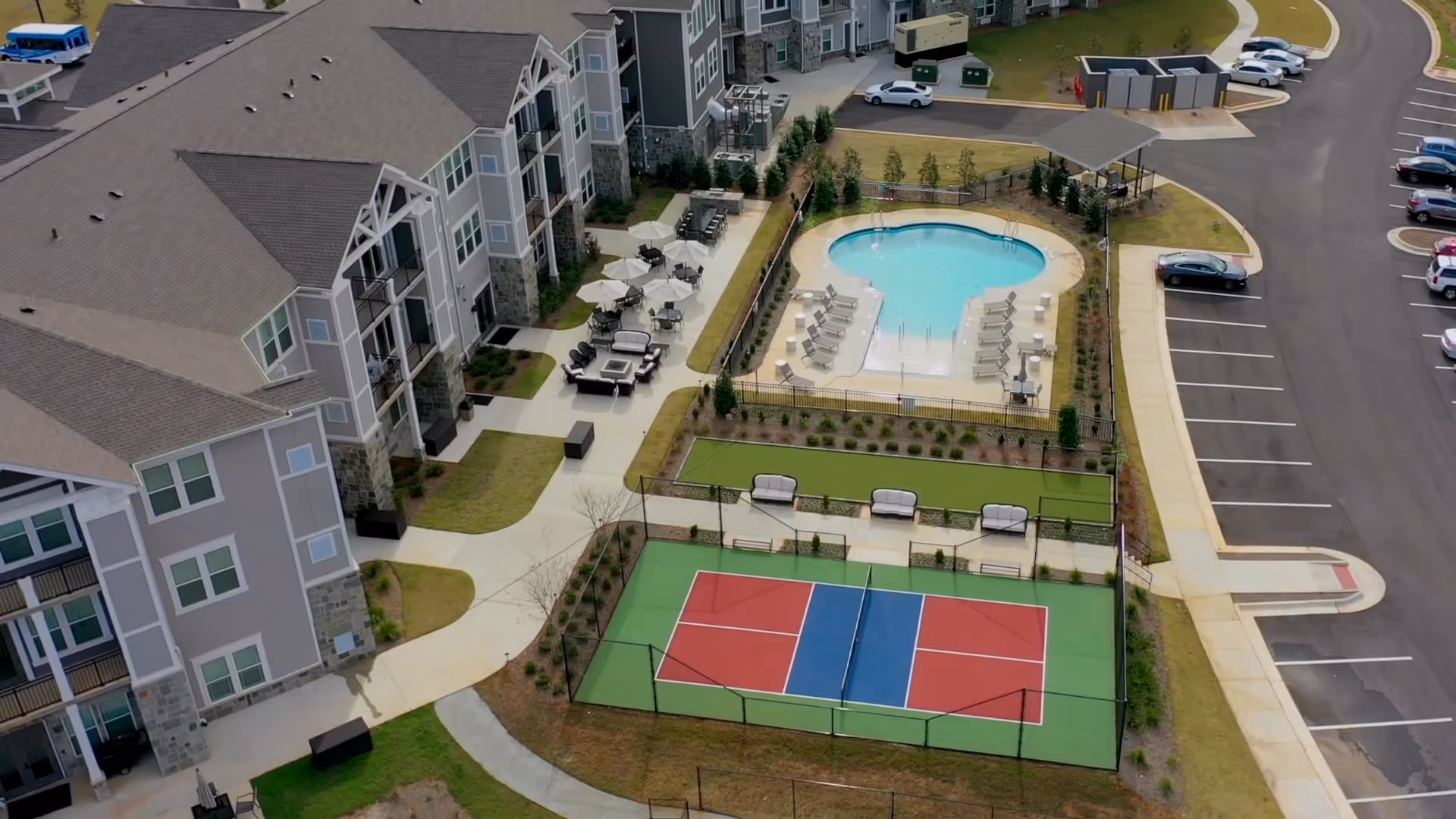 Aerial view of Monark Grove Madison senior living facility showing a multi-story residential building with balconies, an outdoor swimming pool surrounded by lounge chairs and umbrellas, a bocce ball court, a fenced tennis or pickleball court, and a parking lot with several cars.