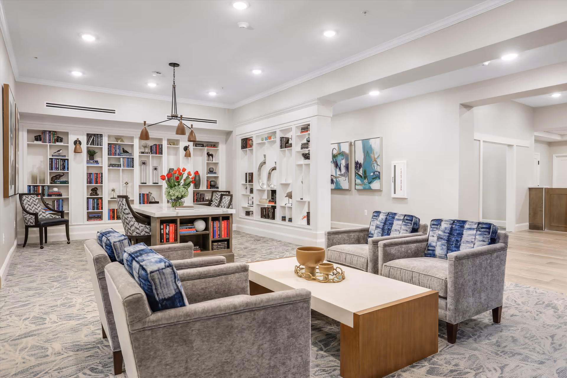 A modern and cozy common area in a senior living facility featuring four gray armchairs with blue patterned cushions arranged around a rectangular coffee table with decorative items. In the background, there is a large built-in bookshelf filled with books and decorative objects, a table with chairs, and two abstract paintings on the wall. The room is well-lit with recessed ceiling lights and has a light-colored carpet with a subtle pattern.