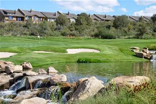 A scenic outdoor view featuring a small waterfall flowing over rocks into a pond, with a well-maintained green golf course and sand bunkers in the background. Residential buildings are visible beyond the golf course under a partly cloudy sky.