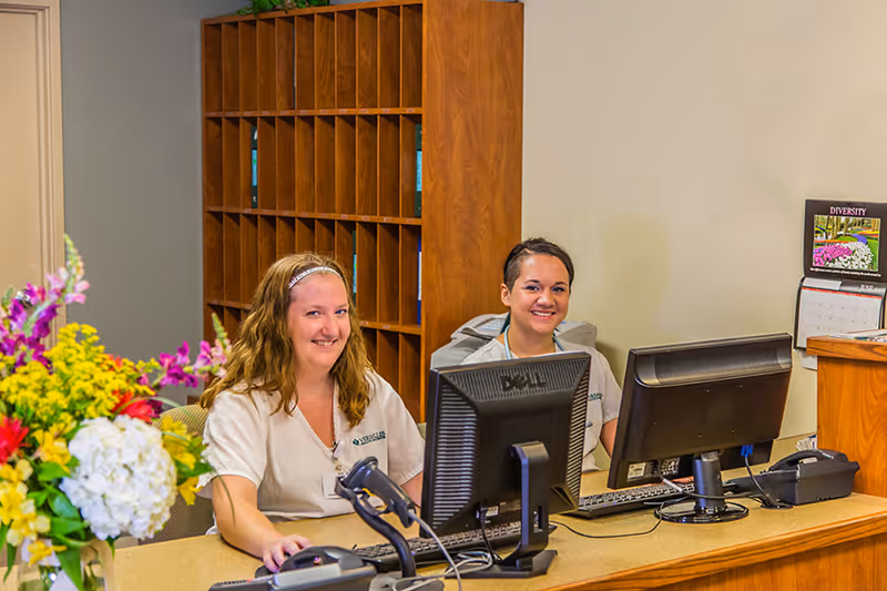 Two smiling female staff members sitting behind a reception desk with computer monitors in a healthcare facility. There is a wooden mail organizer behind them and a colorful flower arrangement on the desk.