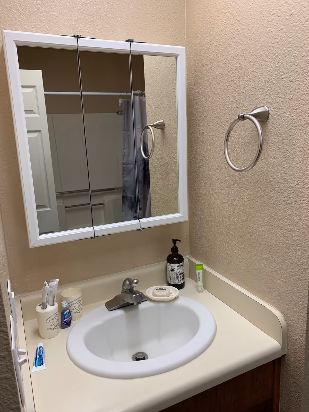 A bathroom sink with a white countertop and a silver faucet. Above the sink is a white-framed mirror cabinet. On the countertop, there are various toiletries including a toothbrush holder with toothbrushes, a small tube of toothpaste, a bottle of hand soap, a bar of soap on a dish, and a small tube of cream. A silver towel ring is mounted on the beige textured wall to the right of the sink.