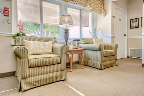 A cozy sitting area with two striped armchairs separated by a wooden side table holding a lamp and decorative items. Behind the chairs is a large window with a beige valance, allowing natural light to brighten the room. The walls are light-colored with white trim, and there is a framed picture and a door visible in the background.