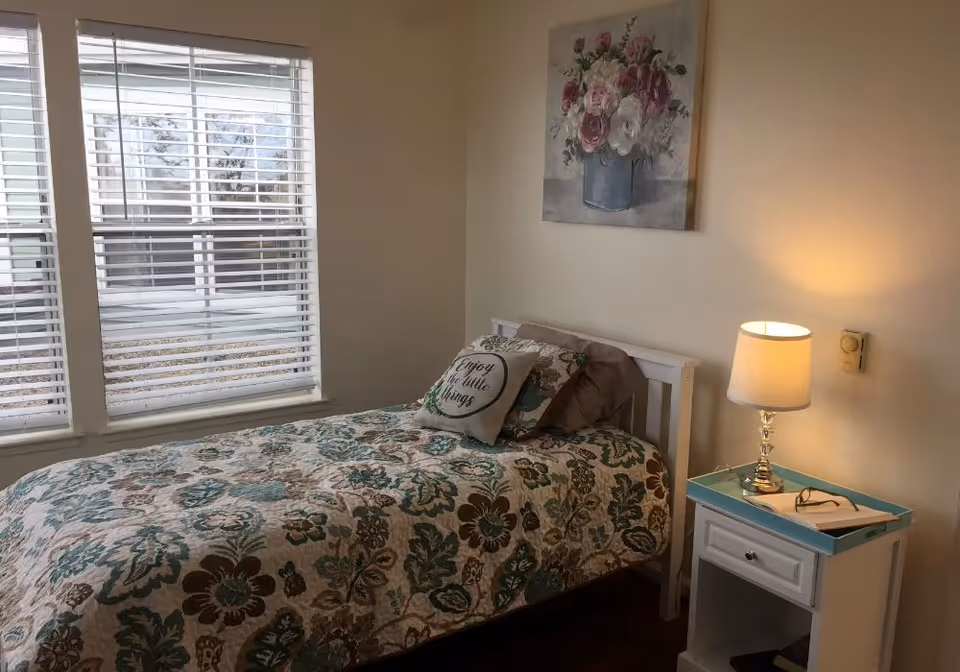 Small bedroom with a single bed covered in a floral quilt, a decorative pillow, a nightstand with a lamp and book, and windows with blinds.
