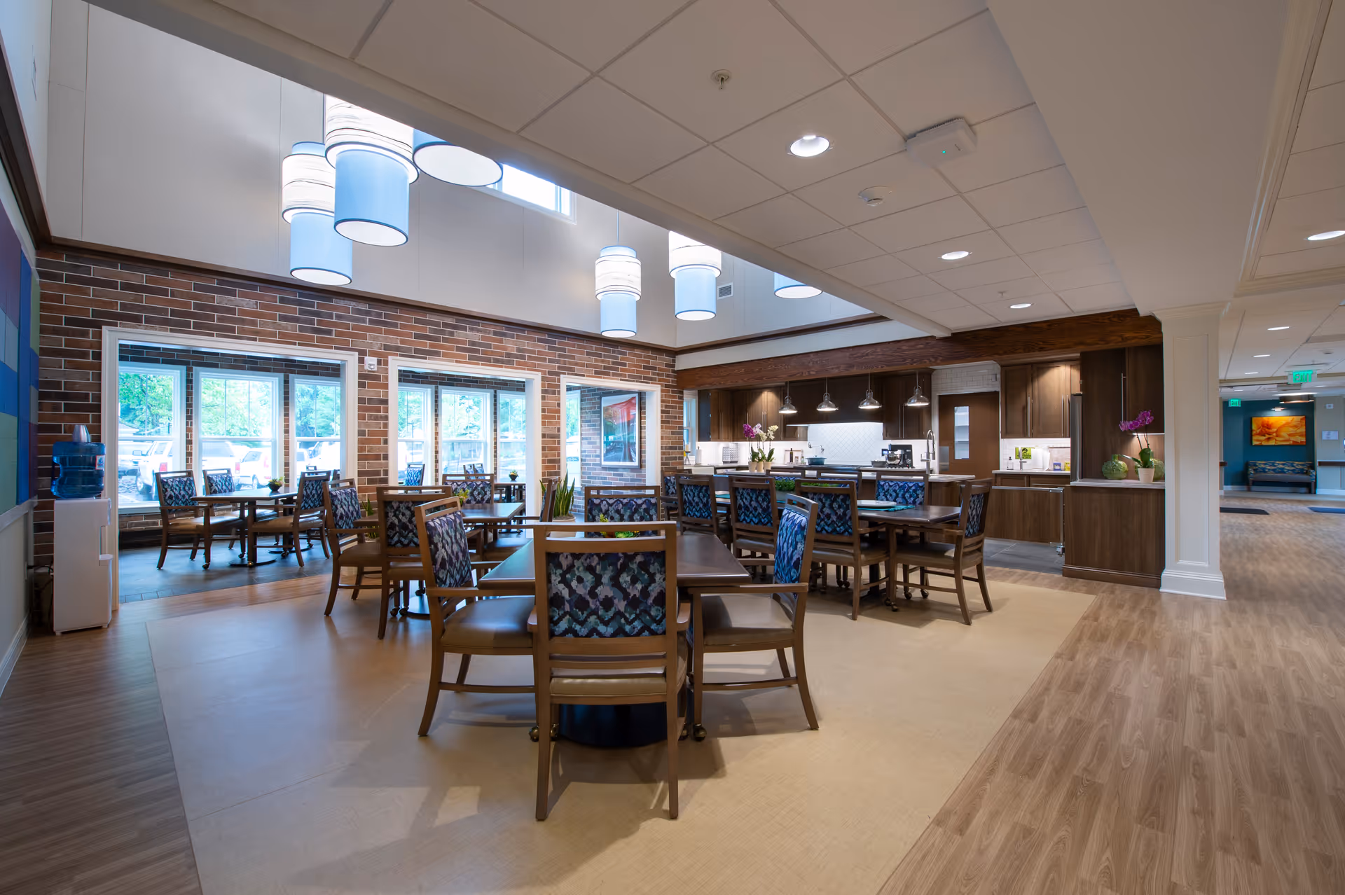 A spacious dining area in a senior living facility with multiple wooden tables and chairs arranged neatly. The room features large windows letting in natural light, brick accent walls, modern pendant lighting, and a kitchen area with dark wood cabinetry and countertops in the background.