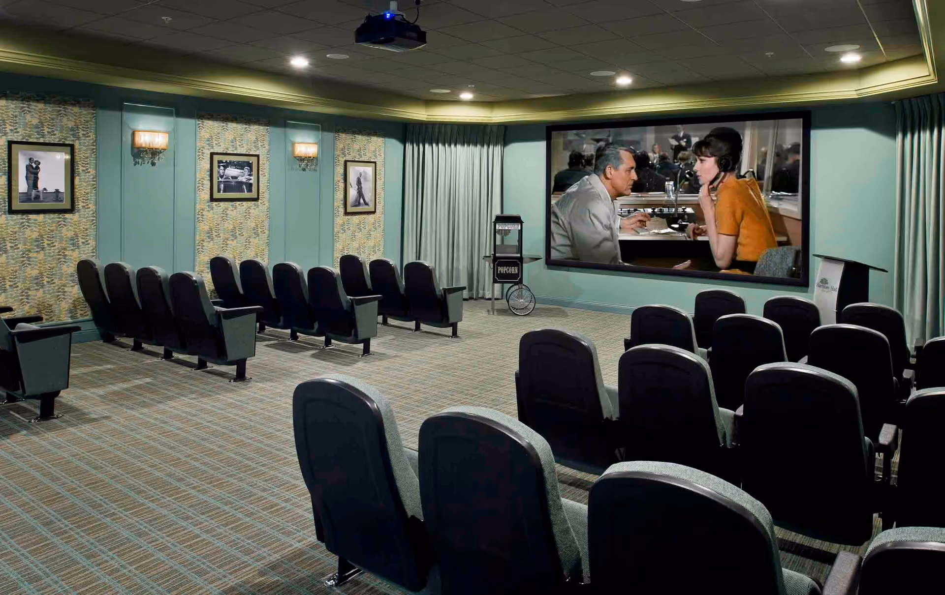 A small movie theater room with rows of black cushioned seats facing a large screen showing a scene from a classic film. The room has patterned carpet, teal walls with framed black and white photos, wall sconces, and a popcorn machine near the screen.