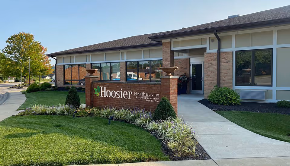 Exterior view of Hoosier Health & Living facility showing a brick sign with the facility name in front of a single-story building with large windows, surrounded by well-maintained landscaping and a sidewalk leading to the entrance.