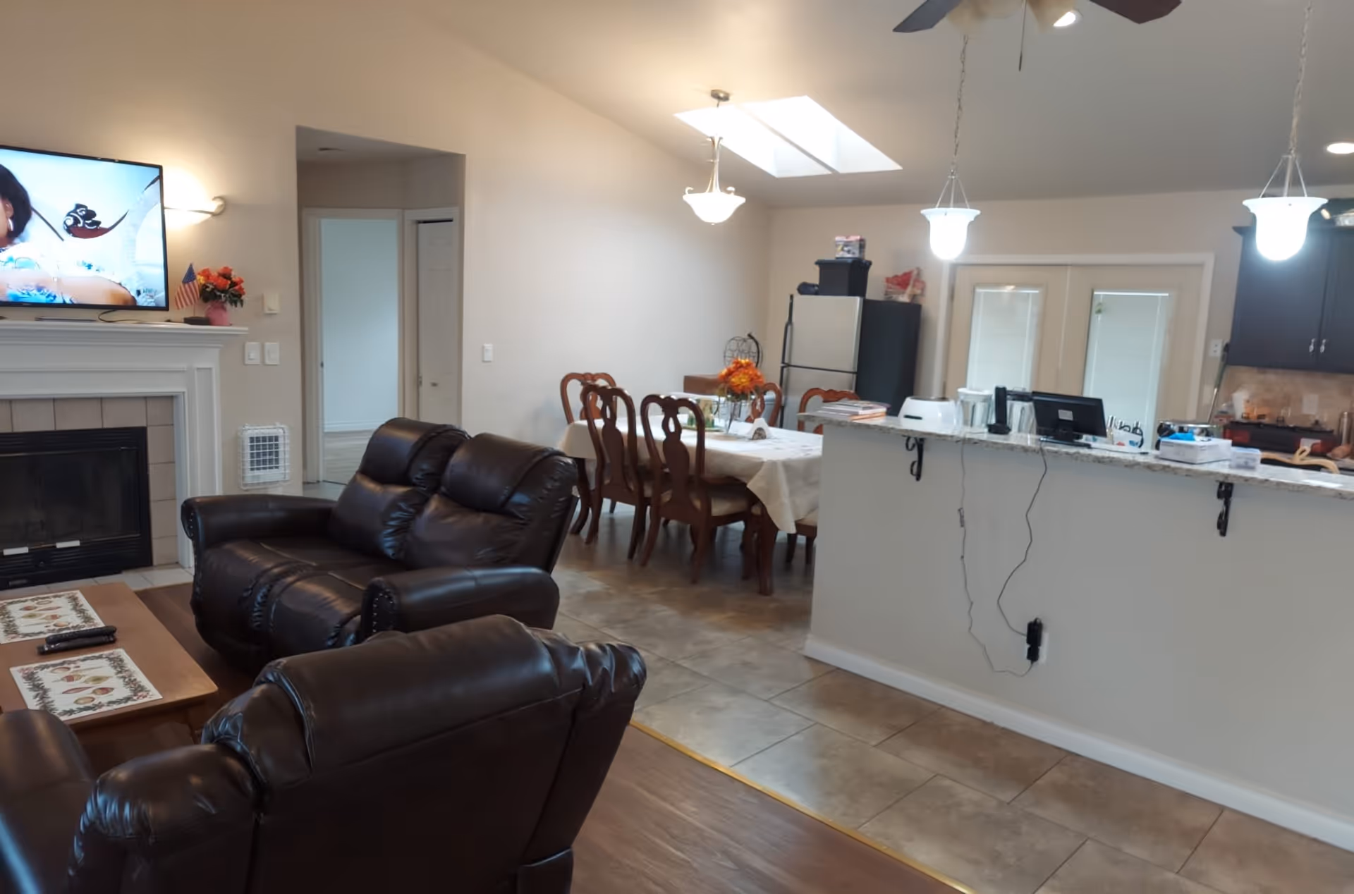 Open-plan living area with dark leather recliners and a fireplace in the foreground, and a dining table and kitchen counter in the background.
