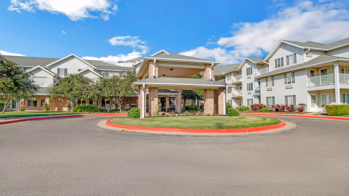 Front entrance and circular drive of a senior living complex with a covered porte-cochere, landscaped roundabout and multi-story residential buildings under a blue sky.