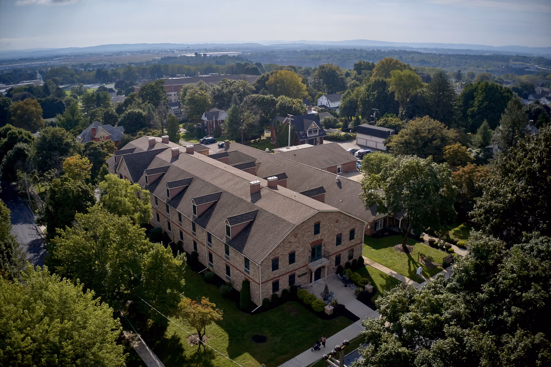 Aerial view of Alexandria Manor, a large brick building surrounded by trees and greenery, with a sidewalk and a few people walking nearby. The background shows a suburban area with more houses and trees extending to the horizon.