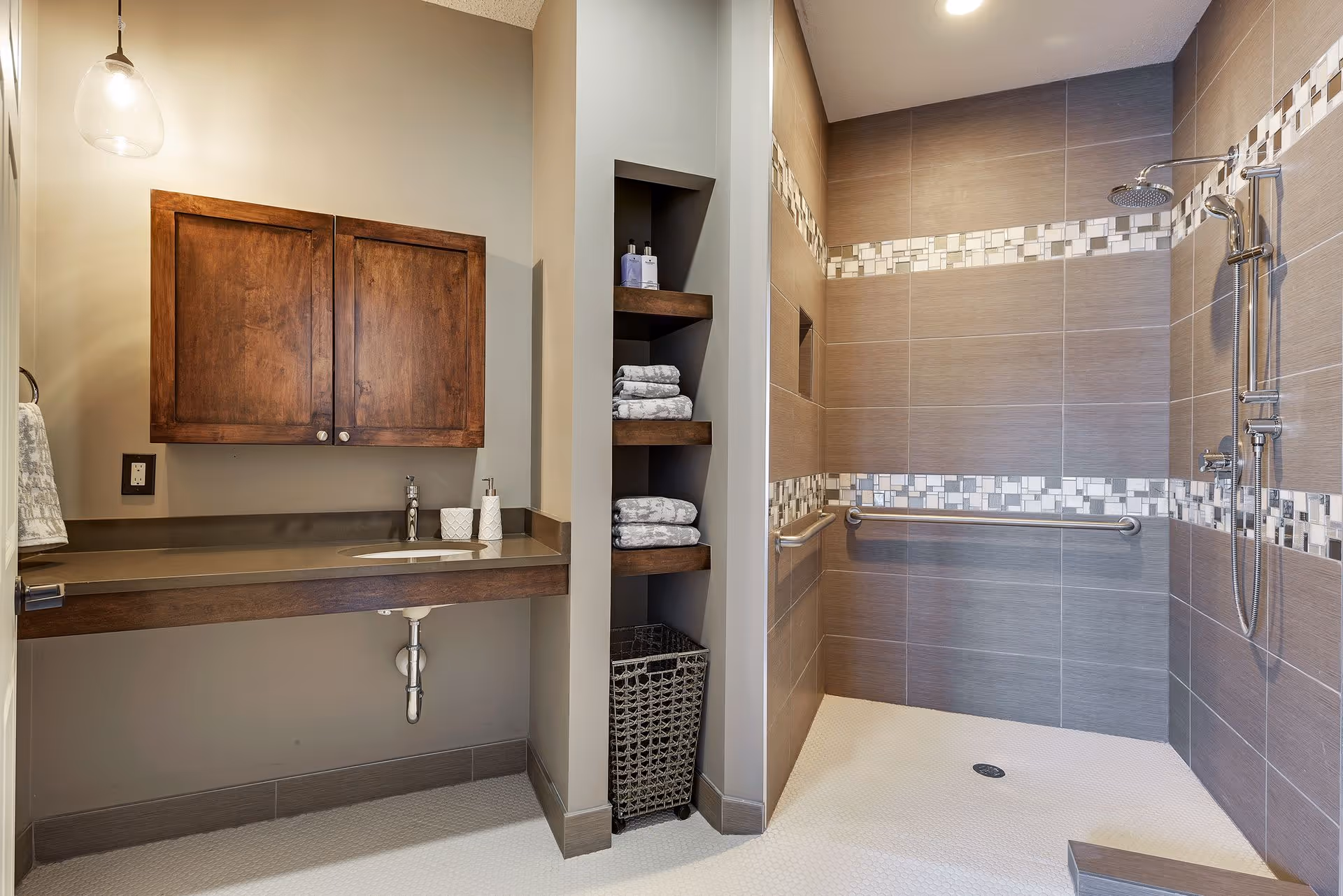 A modern bathroom featuring a walk-in shower with gray tiles and decorative mosaic tile accents. The shower has a rain showerhead, a handheld showerhead, and a grab bar. Next to the shower is a built-in shelving unit with neatly folded towels and toiletries. On the left side, there is a wooden vanity with a sink, soap dispenser, and a wall-mounted cabinet above it. A pendant light hangs from the ceiling, illuminating the space.