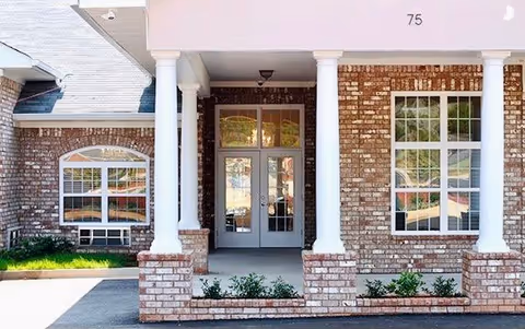 Front entrance of a brick building with white columns, double glass doors, and large windows on either side. The building number 75 is visible above the entrance.