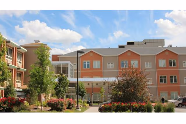 Front exterior of a multi-story senior living building with a landscaped walkway, trees, and entrance under a blue sky.