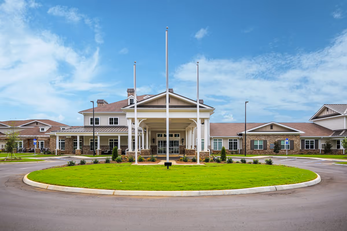 Front exterior view of Grand South Senior Living facility showing a large building with a covered entrance, three flagpoles, well-maintained green lawn, and a circular driveway under a partly cloudy blue sky.