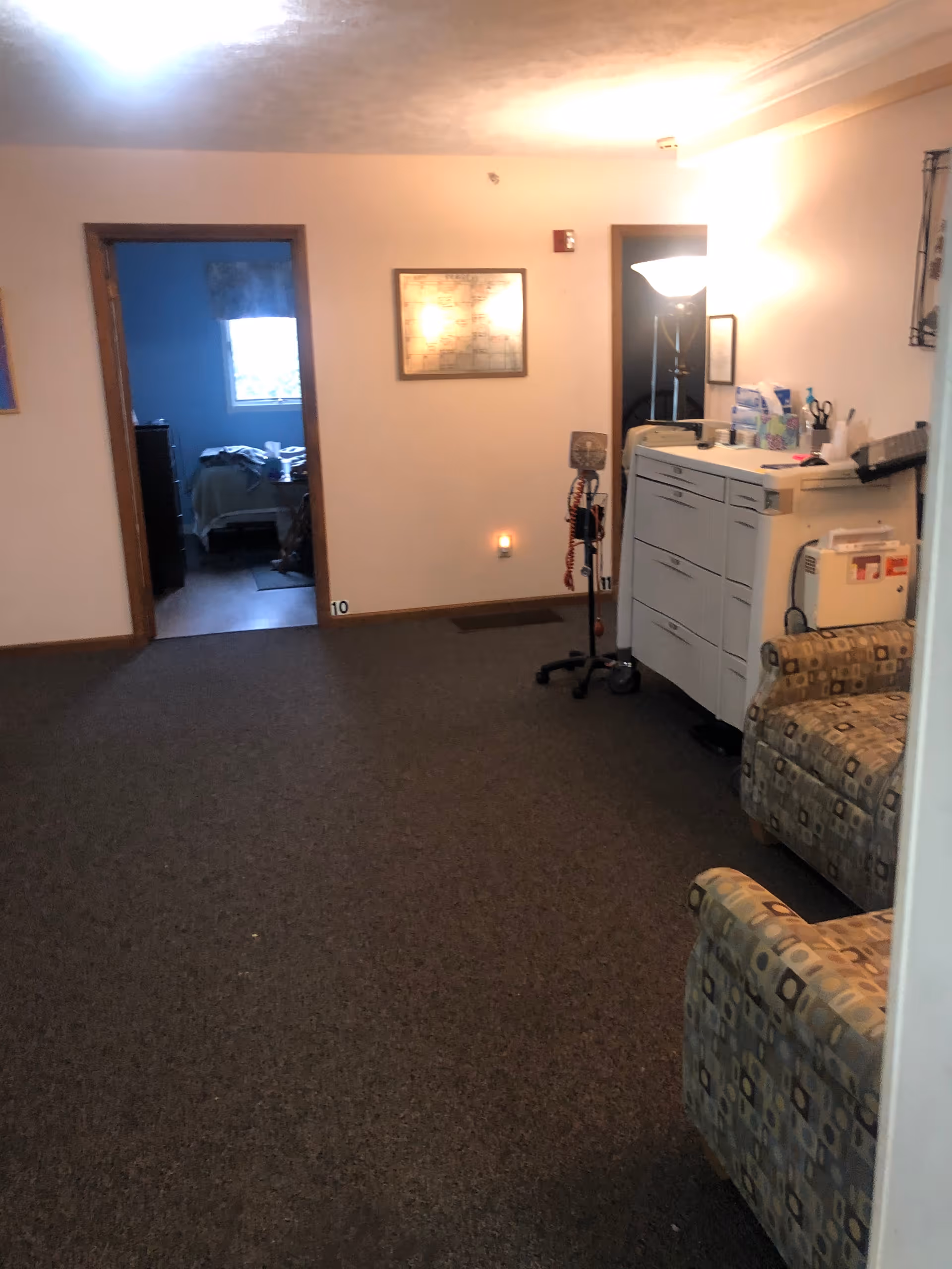 Interior view of a hallway in an assisted living facility with carpeted floor, two patterned armchairs on the right, a white cabinet with medical supplies and a floor lamp. There are two doorways visible, one leading to a bedroom with a bed and window, and the other to a darker room. A framed picture hangs on the wall between the doorways.
