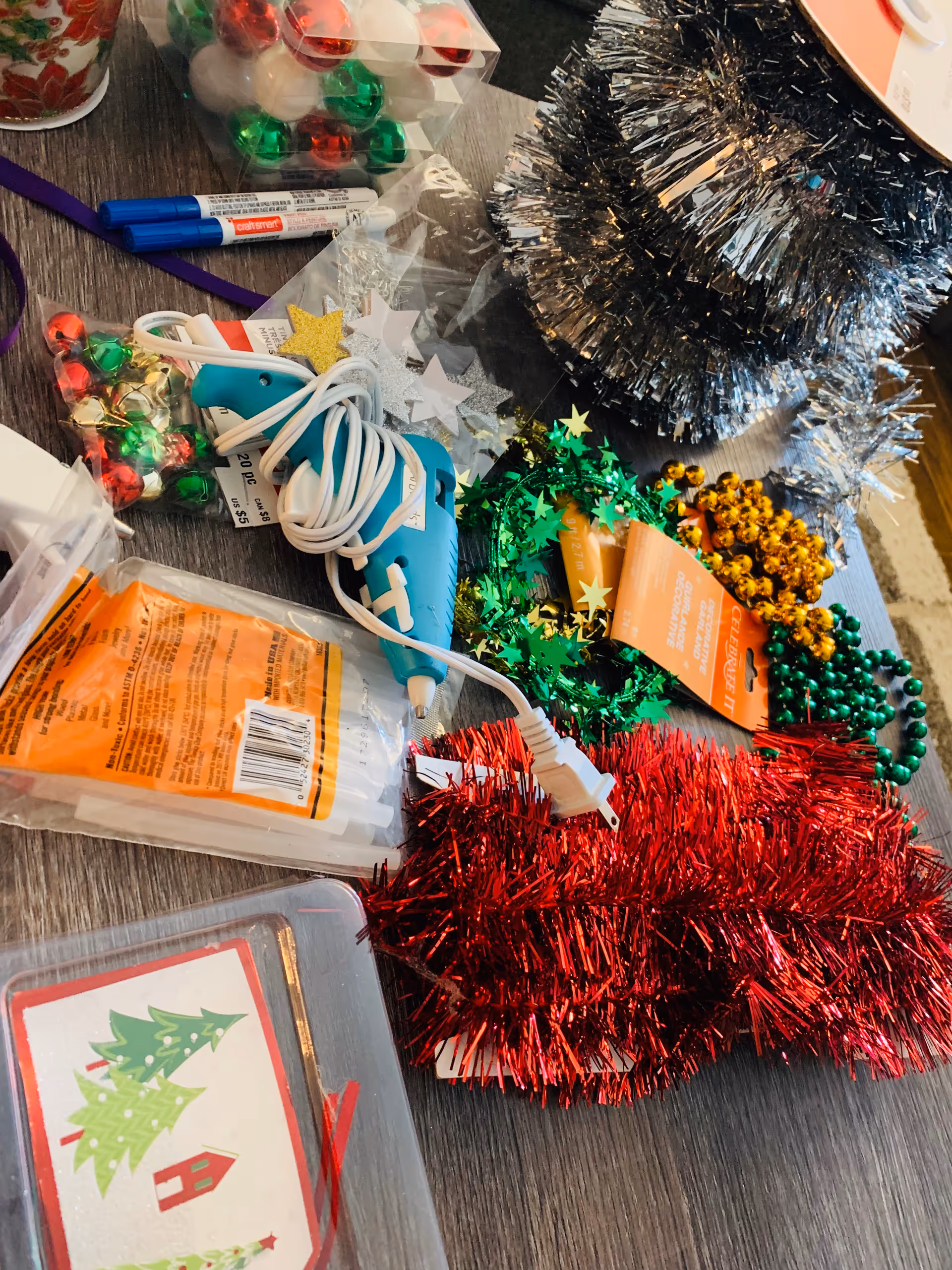 Table covered with Christmas craft supplies including tinsel, beads, a hot glue gun, and ornament balls.