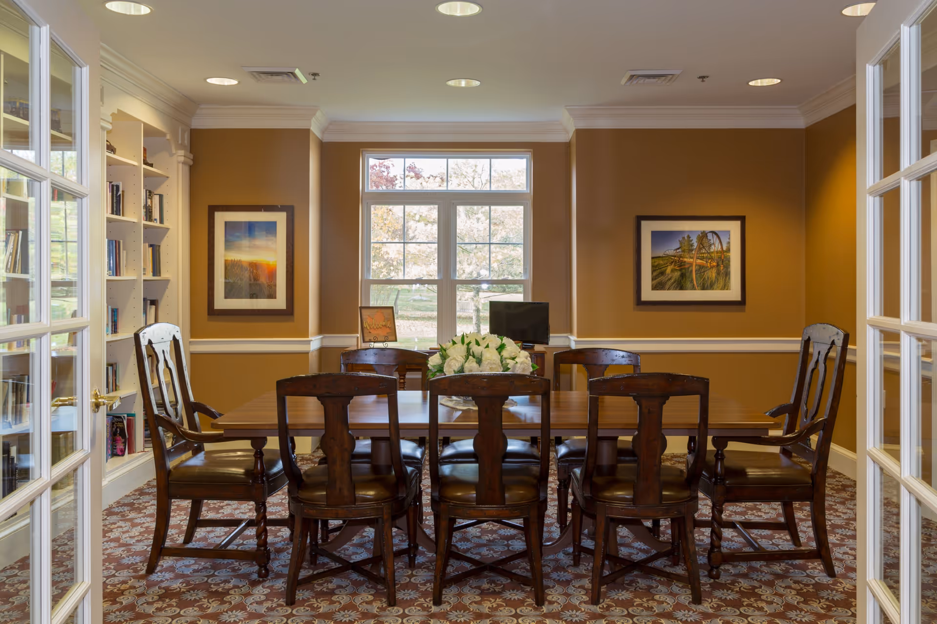 Wooden dining table surrounded by eight chairs in a well-lit meeting/dining room with bookshelves, framed art, and a window.
