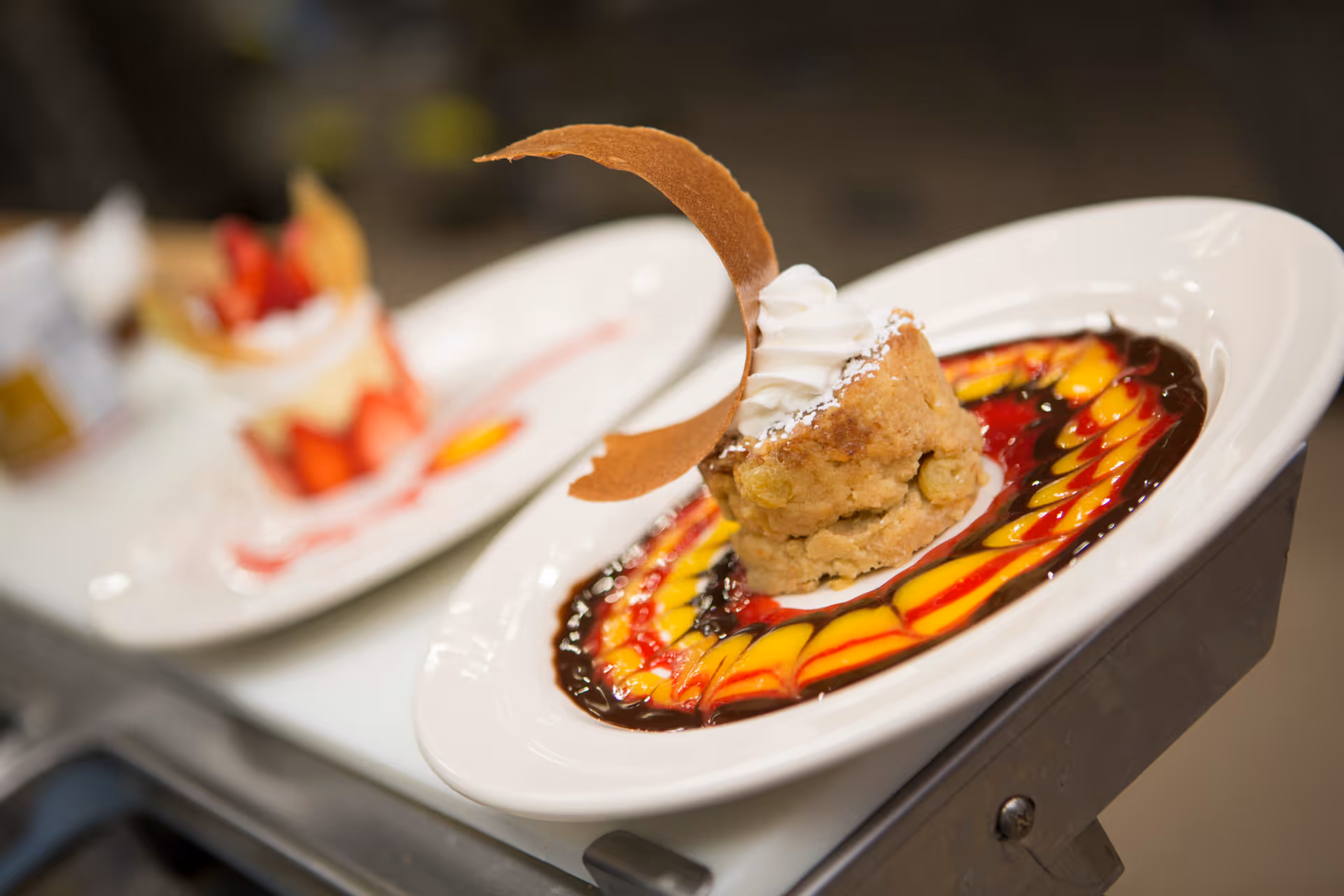 Close-up of a plated dessert with a decorative swirl of chocolate, red, and yellow sauces, topped with whipped cream and a curved cookie garnish. Another dessert plate with strawberries is blurred in the background.