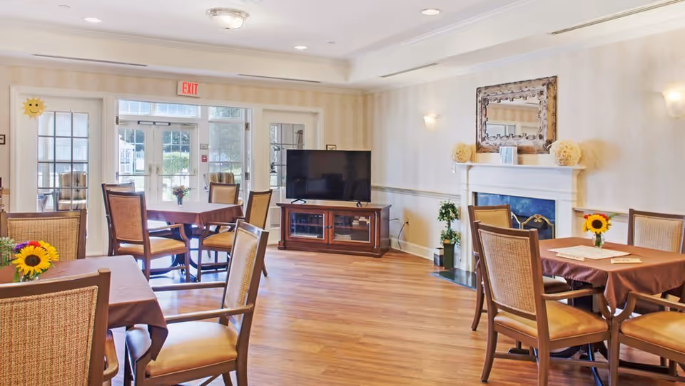A bright and spacious common area with several tables and chairs arranged for dining or socializing. The room features wooden flooring, a large flat-screen TV on a wooden stand, a white fireplace with decorative items on the mantel, and a large mirror above it. Sunflowers in small vases decorate the tables, and large glass doors lead to an outdoor area.