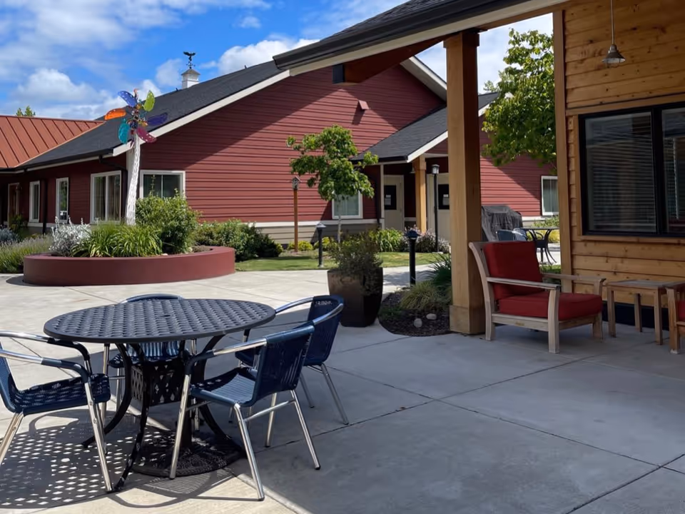 Outdoor patio area with a round metal table and four chairs, a wooden chair with red cushions, and a small wooden side table. In the background, there is a red building with white trim, a colorful pinwheel decoration, green plants, and a partly cloudy blue sky.