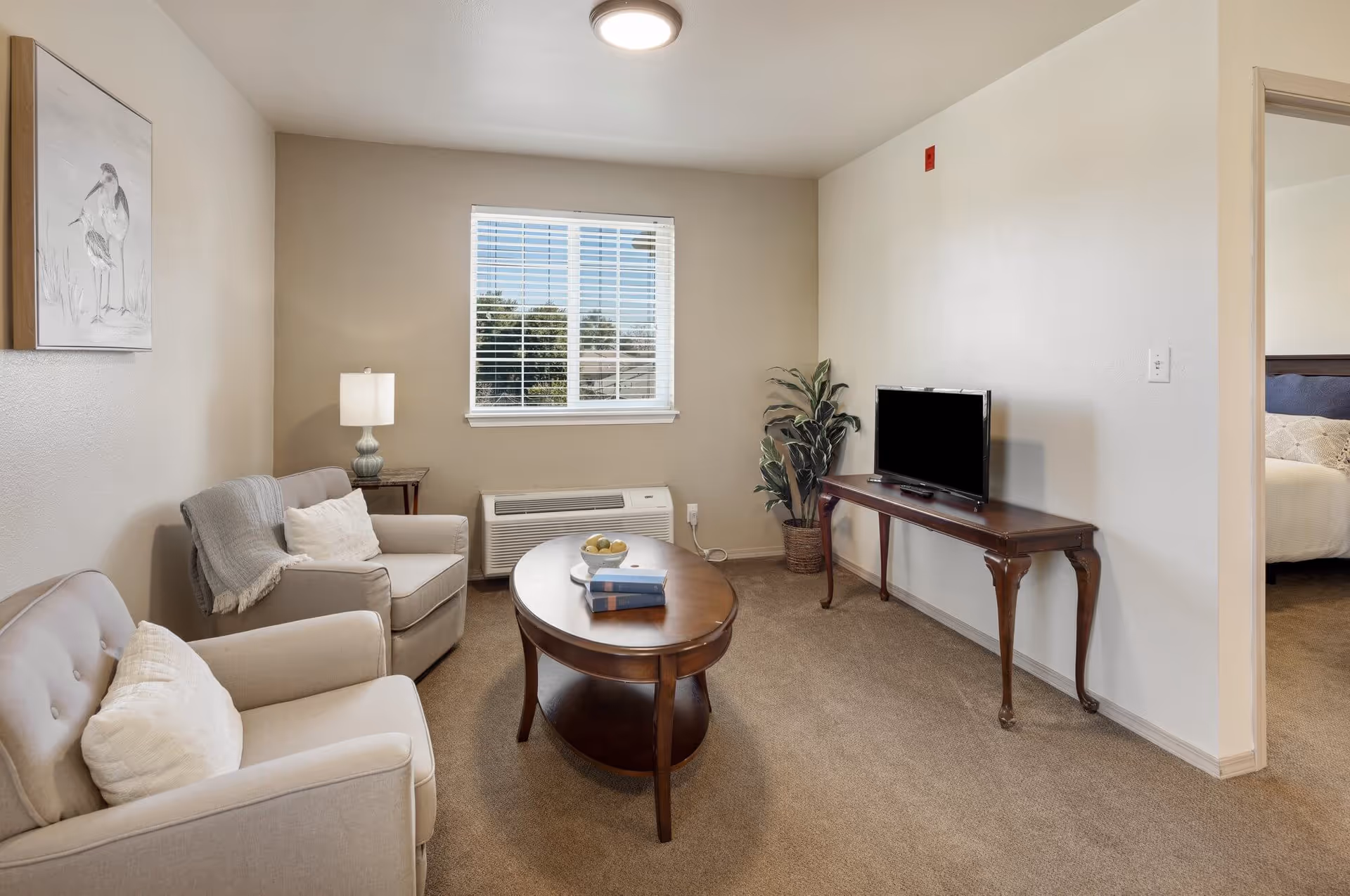 A cozy living room with two beige armchairs, a wooden oval coffee table with books and a bowl of fruit, a wooden TV stand with a flat-screen TV, a potted plant, a window with blinds, a side table with a lamp, and a framed bird artwork on the wall. A bedroom is partially visible through an open doorway.