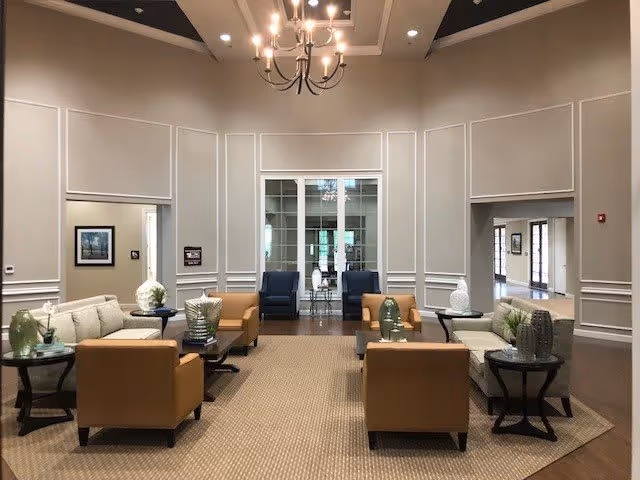 A spacious and elegant living room area with high ceilings and neutral-colored walls. The room features two beige sofas, two tan armchairs, and several small round tables with decorative vases and plants. A chandelier hangs from the ceiling, and large windows and glass doors are visible in the background, allowing natural light to enter the space.
