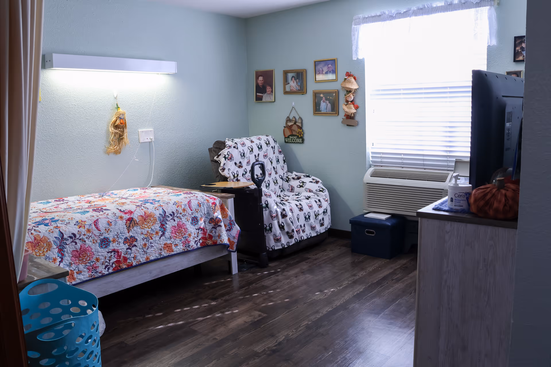 A cozy bedroom in a senior living facility with a single bed covered in a colorful floral quilt, a comfortable armchair draped with a patterned blanket, framed family photos on the wall, a window with blinds and a valance, an air conditioning unit below the window, and a dresser with a television and hand sanitizer on top.