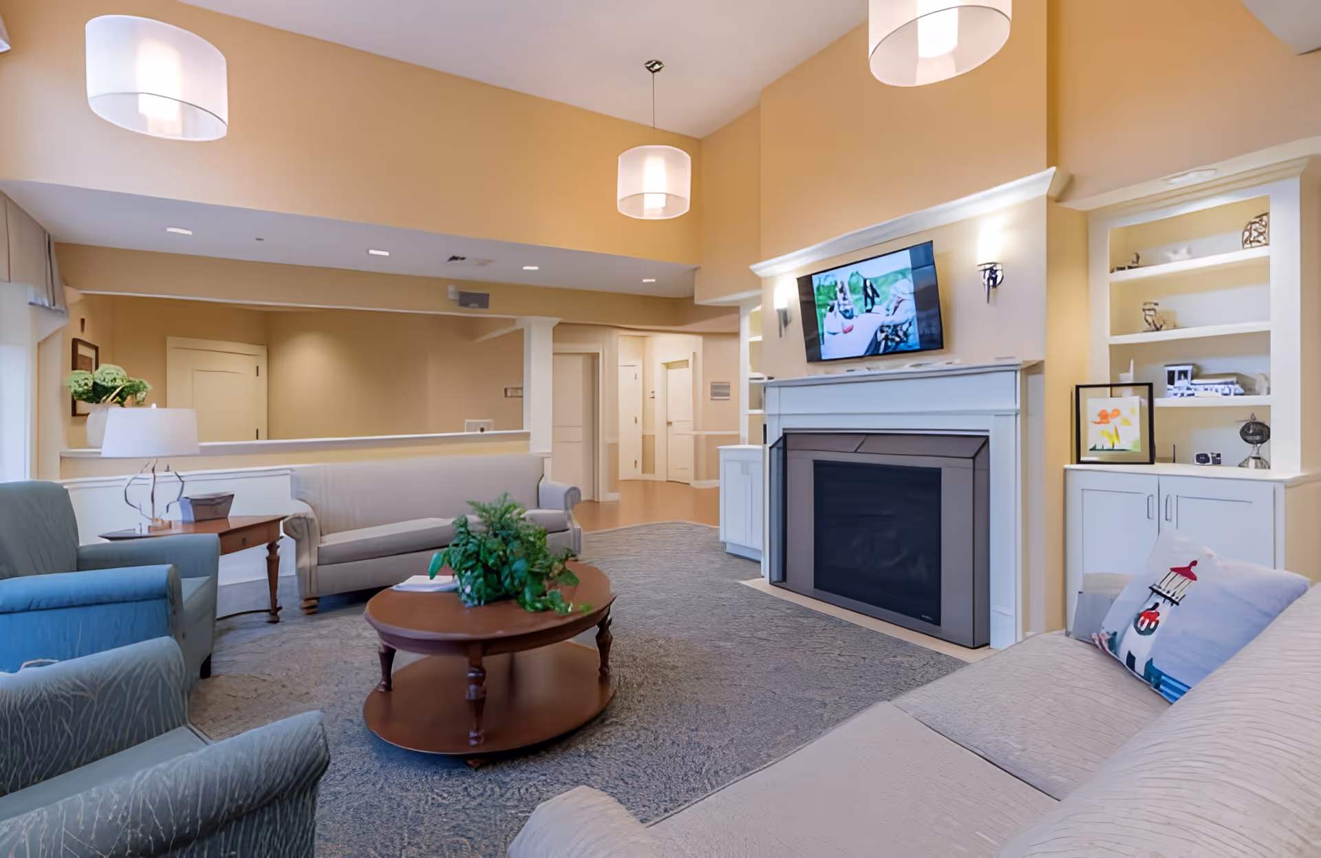 A cozy living room area with beige walls and carpeted floor. The room features a fireplace with a flat-screen TV mounted above it, built-in white shelves with decorative items, a beige sofa with a lighthouse-themed pillow, two blue armchairs, a wooden coffee table with a green plant centerpiece, and a side table with a lamp and plant. Three pendant lights hang from the ceiling.