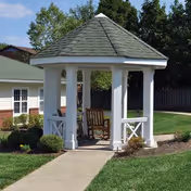 A white gazebo with a gray shingled roof situated on a concrete pathway surrounded by green grass and bushes, with residential buildings and trees in the background.