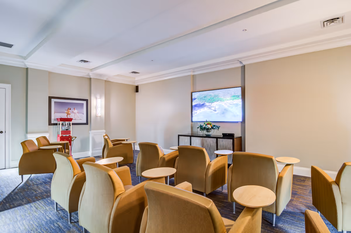 A small theater-style room with several rows of tan armchairs, each with a small round wooden side table. A large flat-screen TV is mounted on the beige wall at the front of the room, above a console table with a flower arrangement. There is a popcorn machine and framed artwork on the left side of the room.