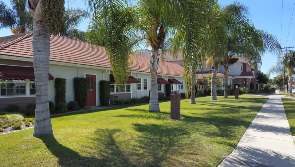 Exterior view of a senior living facility with a row of single-story buildings featuring red doors and awnings, surrounded by palm trees and a well-maintained lawn with a sidewalk running alongside.