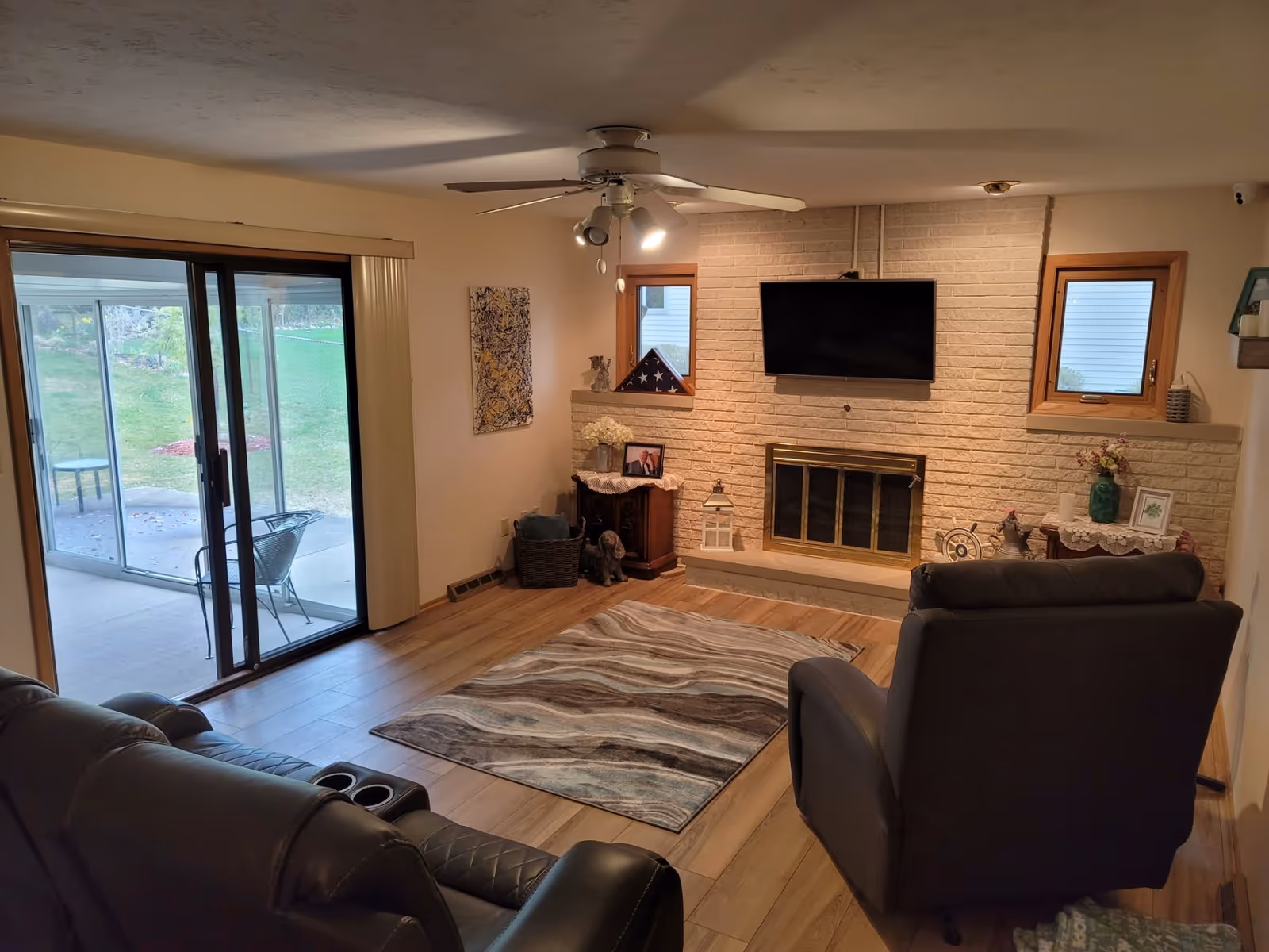 A cozy living room with wooden flooring, a patterned area rug, a ceiling fan with lights, a wall-mounted flat-screen TV above a brick fireplace with a brass screen, and two small windows on either side of the fireplace. There are two dark-colored recliners facing the TV, a sliding glass door leading to an outdoor patio with chairs and a table, and various decorative items including framed photos, flowers, and a folded American flag on a small cabinet.