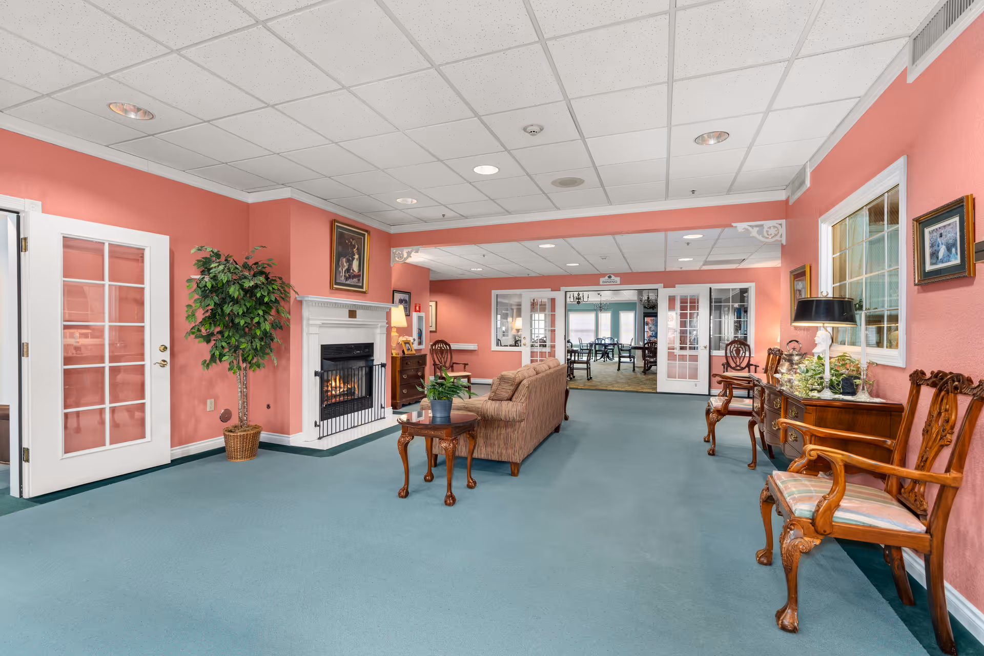 A spacious senior living lounge with sofas, wooden chairs, a fireplace, pink walls and teal carpet.