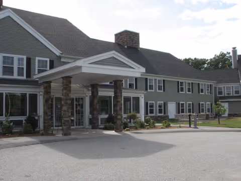 Exterior view of a two-story senior living facility building with gray siding, multiple windows, and a covered entrance supported by stone pillars. There is a paved driveway and some landscaping with small bushes and trees.