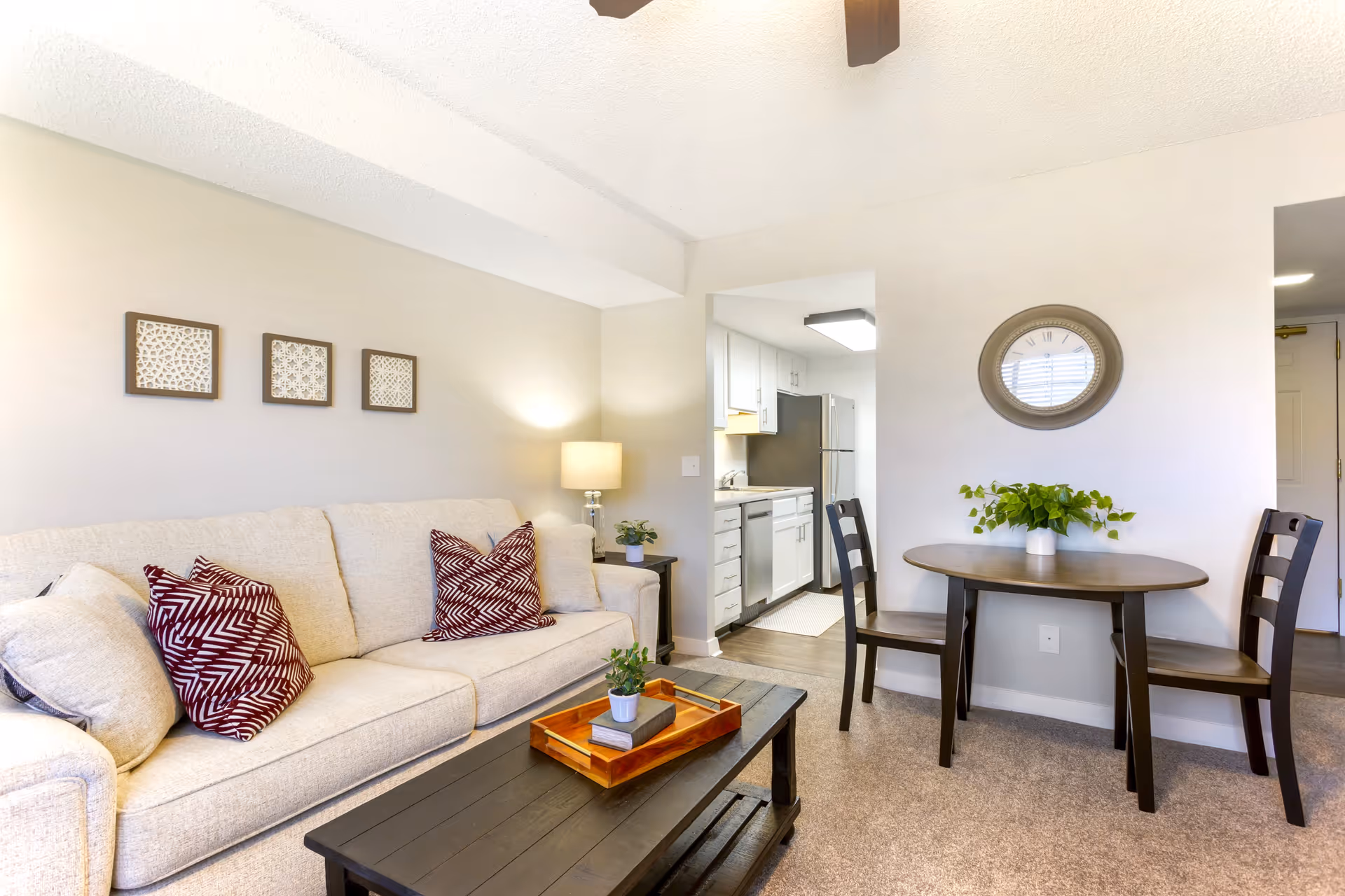A cozy living area in The Montclair Senior Living featuring a beige sofa with patterned red and white pillows, a dark wooden coffee table with a tray and small plant, a small dining table with two chairs and a green plant centerpiece, a wall clock above the dining table, and a view into a kitchen with white cabinets and stainless steel appliances.
