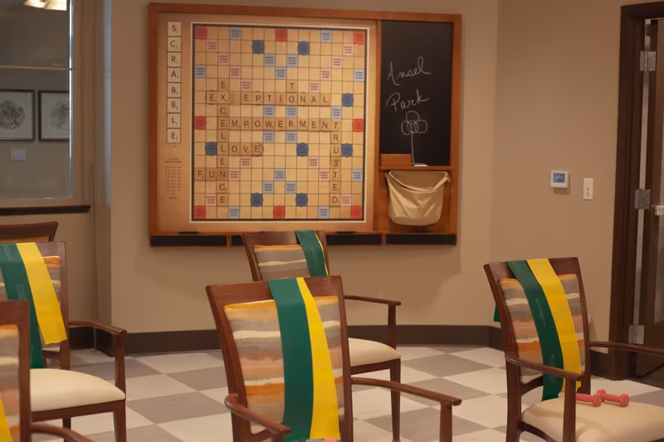 A communal activity room with wooden chairs draped in colorful sashes facing a large wall-mounted Scrabble board and a chalkboard labeled 'Ansel Park'.