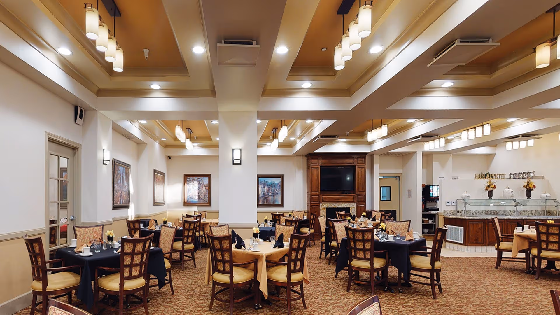 A spacious dining room with multiple tables covered in black and beige tablecloths, each set with napkins, glasses, and small flower arrangements. The room features warm lighting from ceiling fixtures, framed artwork on the walls, and a wooden cabinet with a television in the background. There is also a buffet area with a glass sneeze guard and decorative flowers.