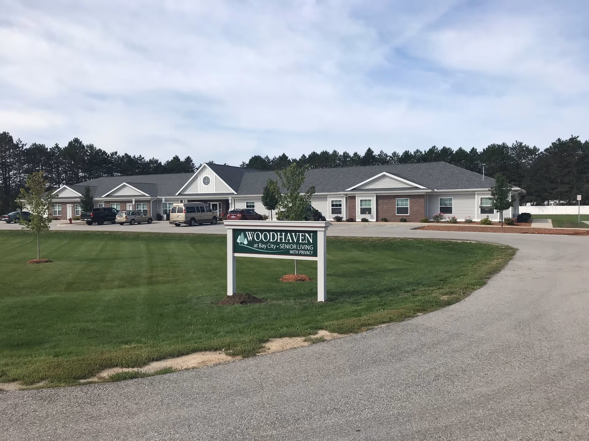 Exterior view of Woodhaven at Bay City senior living facility showing a single-story building with a parking lot and several vehicles. A green sign with white text reading 'Woodhaven at Bay City - Senior Living with Privacy' is prominently displayed on a grassy area near the driveway entrance.