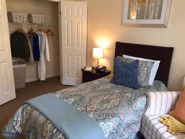 A tidy bedroom with a single bed covered in a patterned bedspread, a nightstand with a lamp, an open closet, and a striped armchair.
