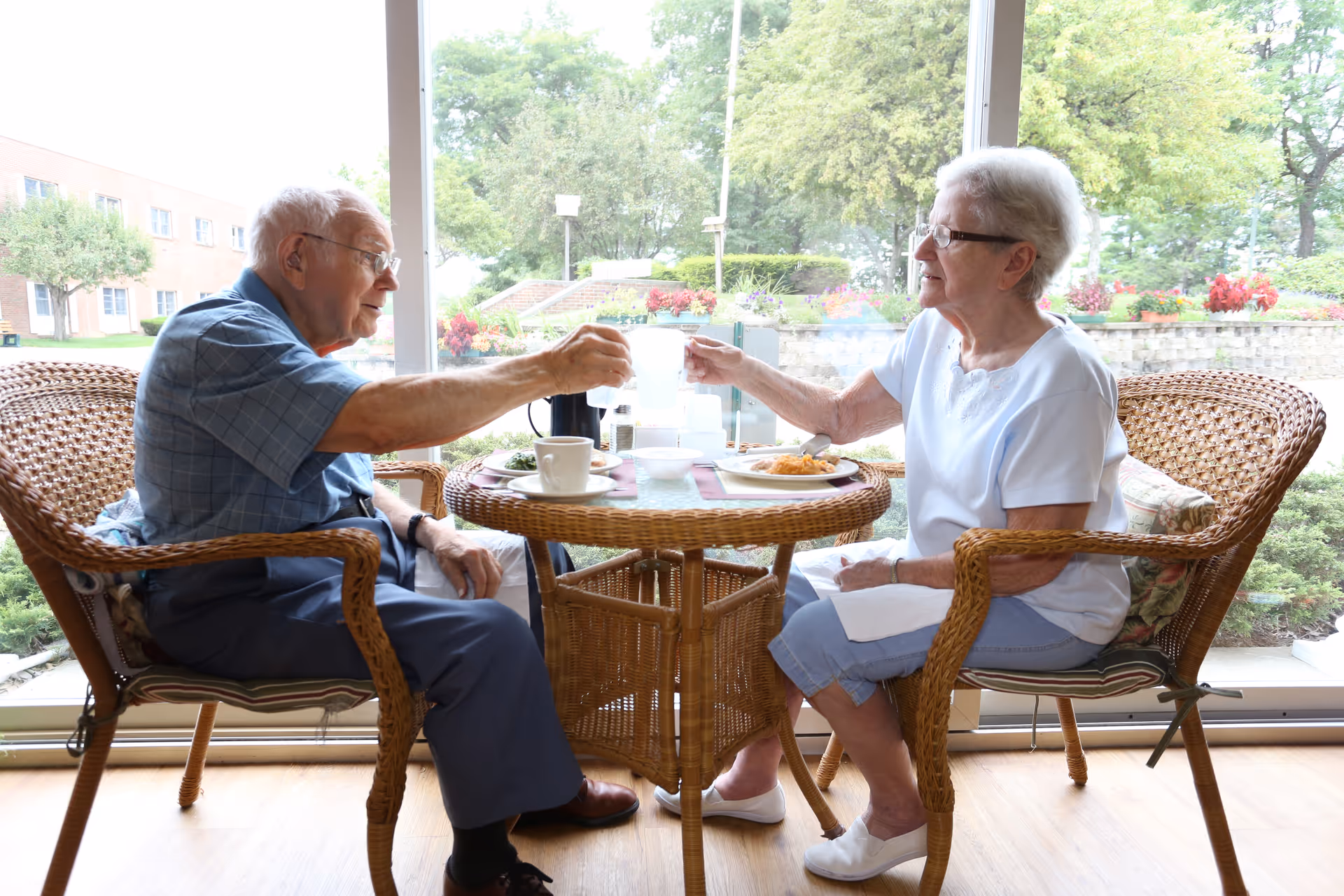 An elderly man and woman sitting at a round wicker table indoors near a large window, toasting with clear glasses. The table has plates of food, cups, and a coffee pot. Outside the window, there is greenery and a building visible.
