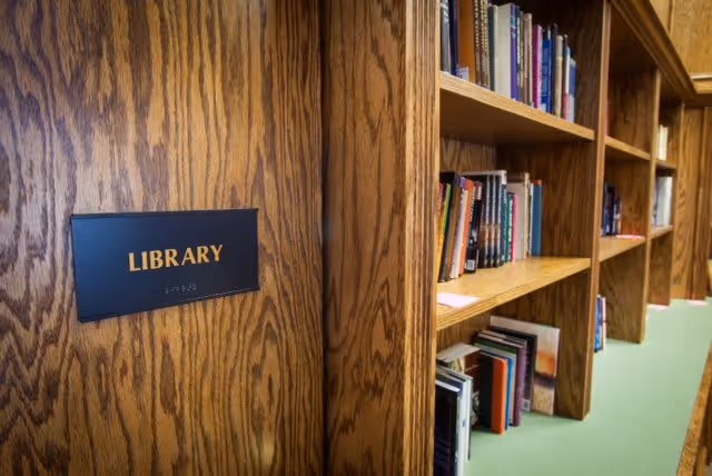Close-up view of a wooden bookshelf filled with books inside a library. A wooden wall panel with a black sign labeled 'LIBRARY' in gold letters is visible on the left side.
